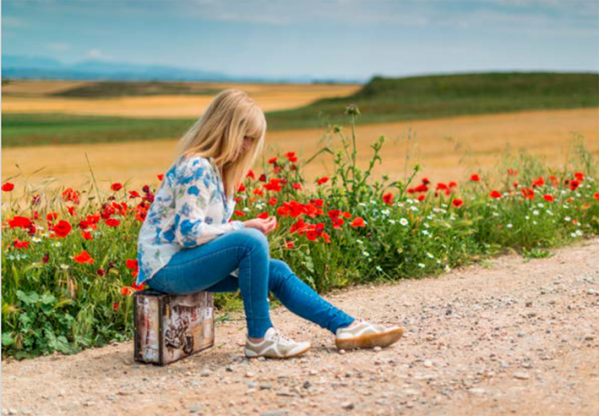 Nainen istuu matkalaukun päällä soratien laidassa, takana punaisia ja valkoisia kukkia ja viljapelto.