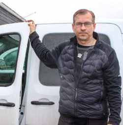 Timo Veijanen seisoo auton vieressä, käsi ovella. Lyhyet hiukset, silmälasit ja tumma takki.