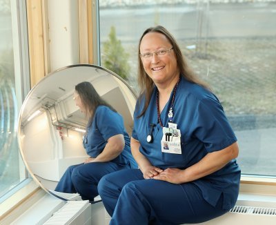 Eva-Maria Strömsholm sinisessä sairaanhoitajan asussa, istuu ikkunalaudalla ja hymyilee. Peilikuva näkyy viereisestä peilistä. 