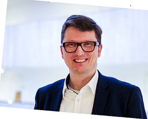 Jukka Mikkonen valkoisessa kauluspaidassa ja tummassa takissa, tummasankaiset silmälasit, tummat hiukset, hymyilee.