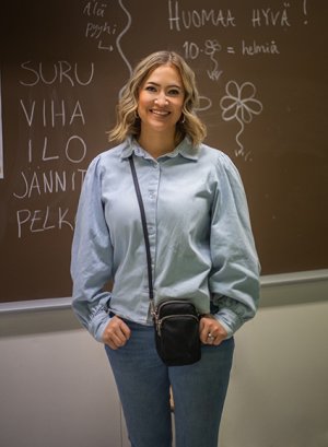 Ella Saarenpää-Kervinen seisoo sinisessä paidassa ja farkuissa liitutaulun edessä, johon on kirjoitettu tekstiä valkoisella liidulla.
