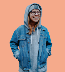 Iloinen silmälasipäinen nuori nainen pukeutuneena harmaaseen huppariin, siniseen farkkutakkiin seisoo oranssin seinän edessä harmaa pipo ja hupparin huppu päässä.