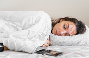 Tummahiuksinen nainen nukkuu valkoinen tyyny pään alla ja valkoiseen peittoon kääräytyneenä.