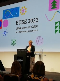 Henkilö seisoo puhujakorokkeen takana, taustalla iso kuva heijastettuna, tekstit EUSE 2022 June 20-22 Oslo.